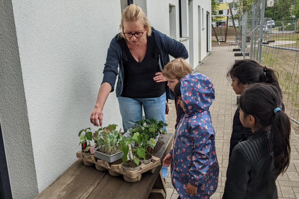 Eine erwachsene Person zeigt einer Gruppe von Kindern verschiedene Pflanzen auf einem Tisch.