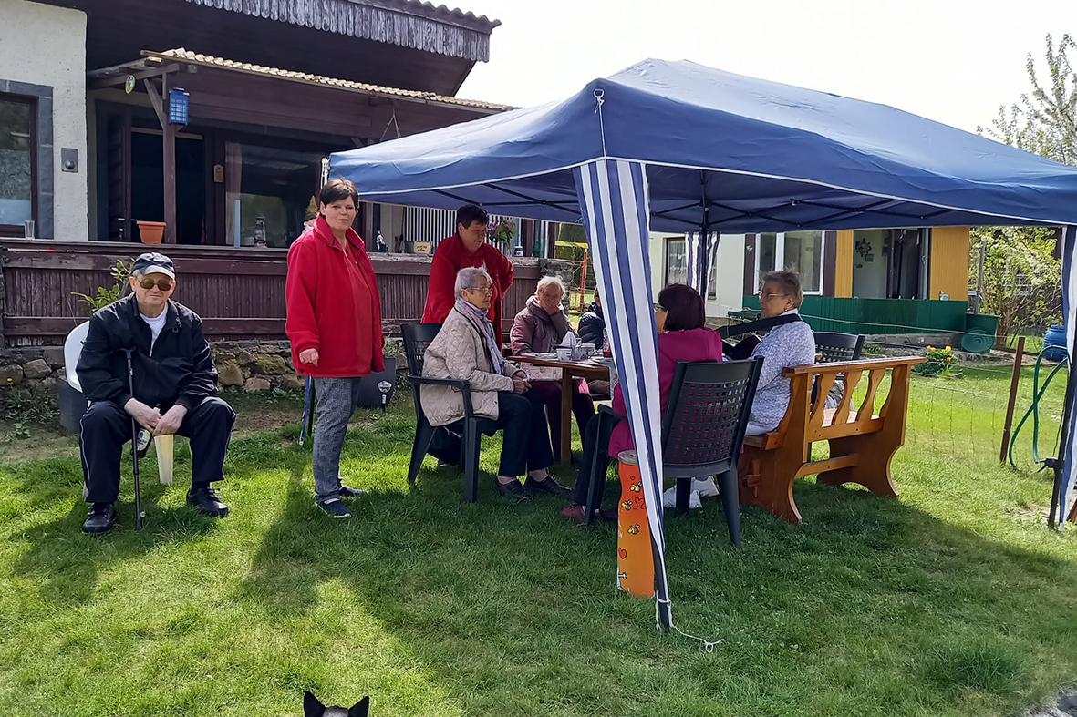 Eine Gruppe von Menschen sitzt und steht unter einem blauen Pavillon auf einem Rasen vor einem Haus.