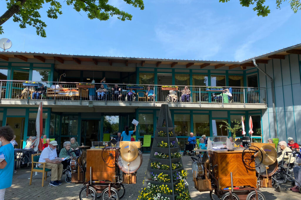 Menschen versammeln sich vor einem Gebäude mit Balkon bei sonnigem Wetter.