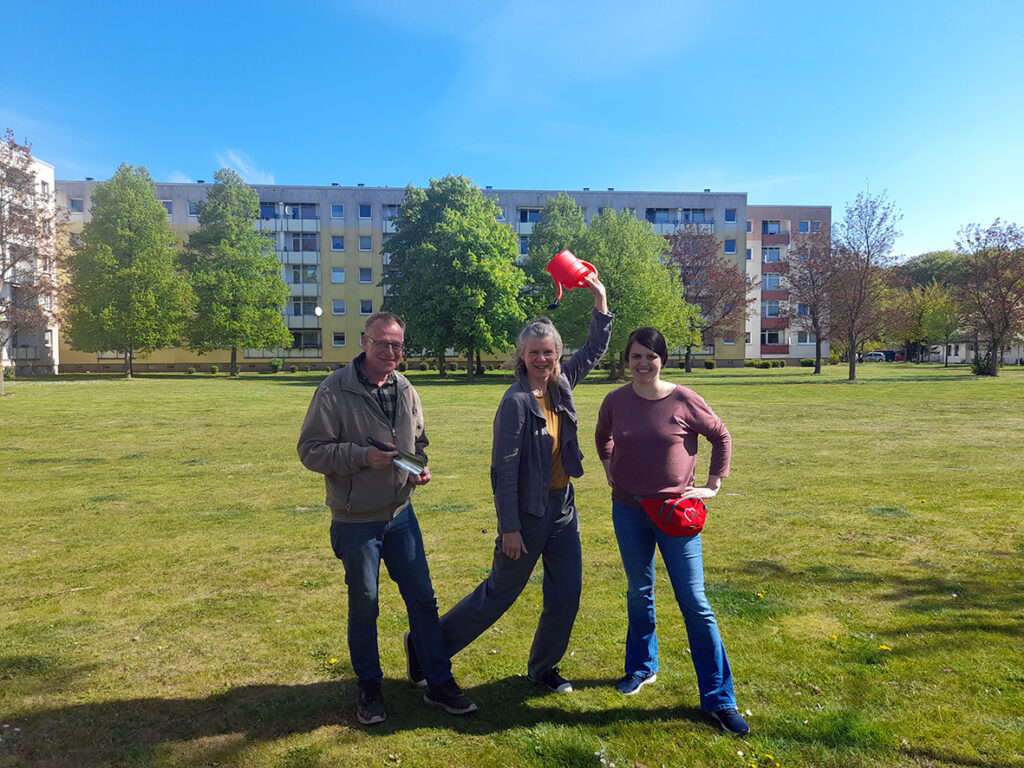 Drei Personen stehen draußen auf einer Wiese mit einer Gießkanne.