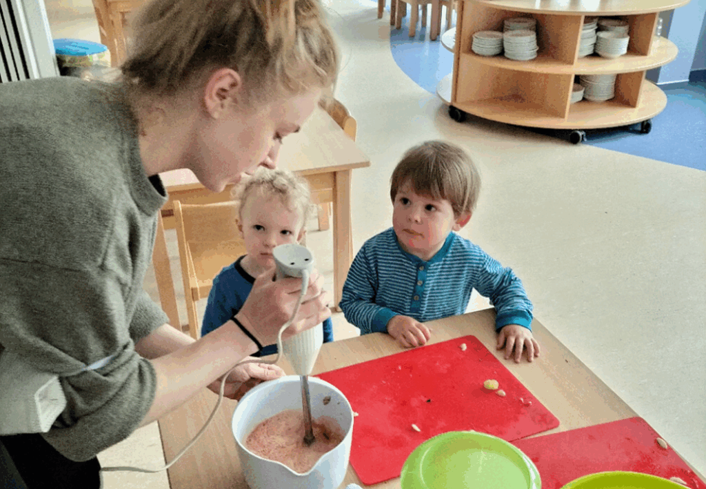 Eine erwachsene Person mit einem Stabmixer und zwei Kinder am Tisch
