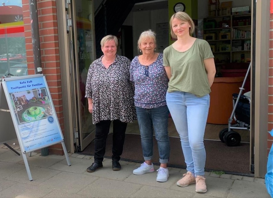 Drei Frauen stehen vor einem Gebäude neben einem Schild mit der Aufschrift 'Café F.u.N. Treffpunkt für Familien'.