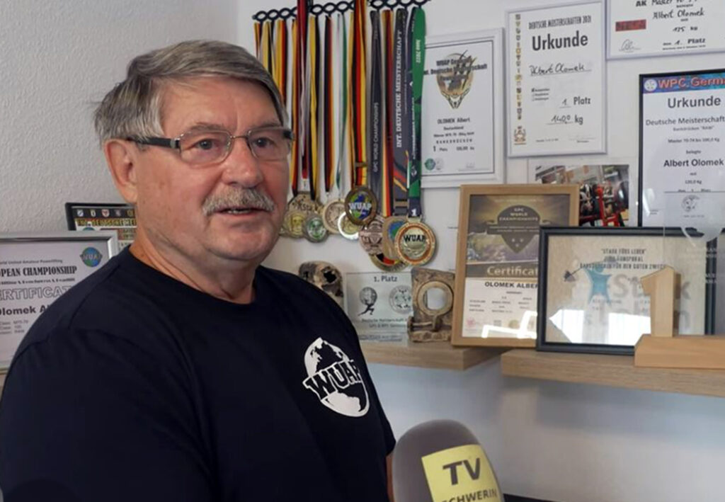 Ein Mann steht vor einer Wand mit Medaillen und Urkunden.