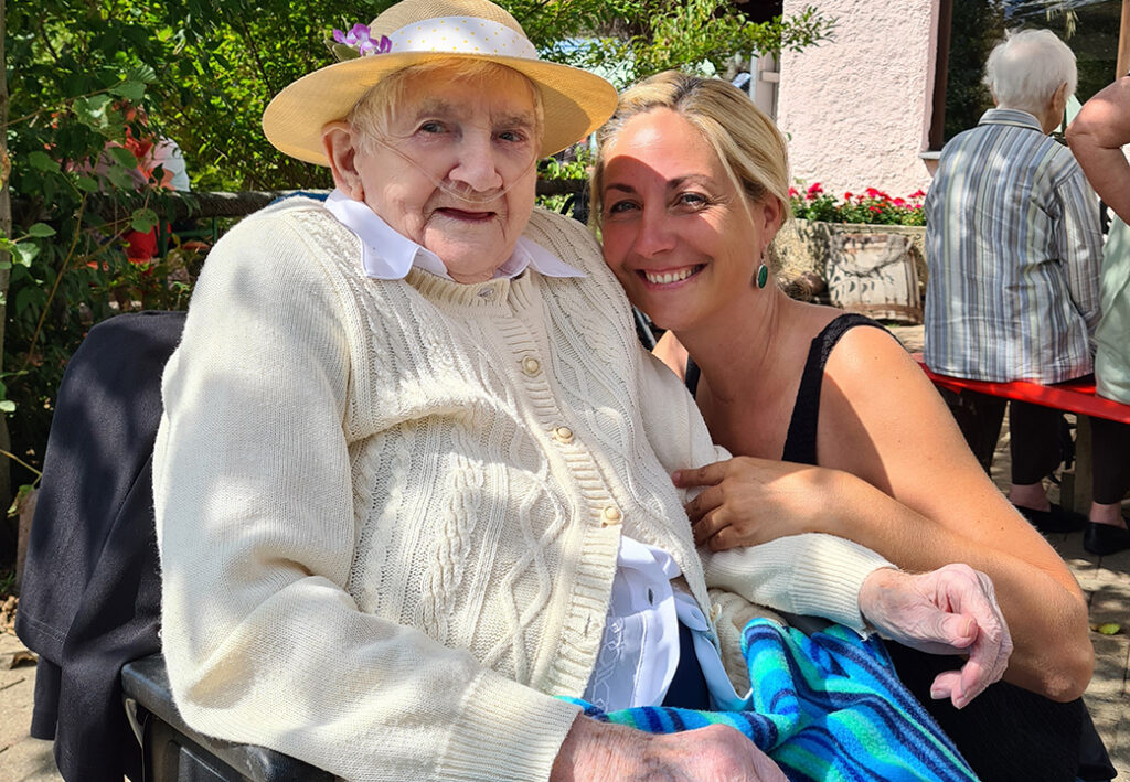 &Auml;ltere Frau in Strickjacke und Hut neben einer l&auml;chelnden jungen Frau im Freien.