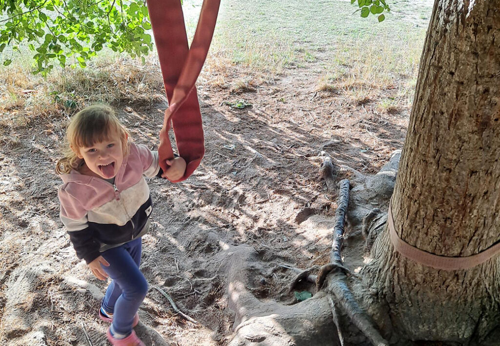 Ein lachendes Kind h&auml;lt eine Slackline, die an einem Baum befestigt ist.