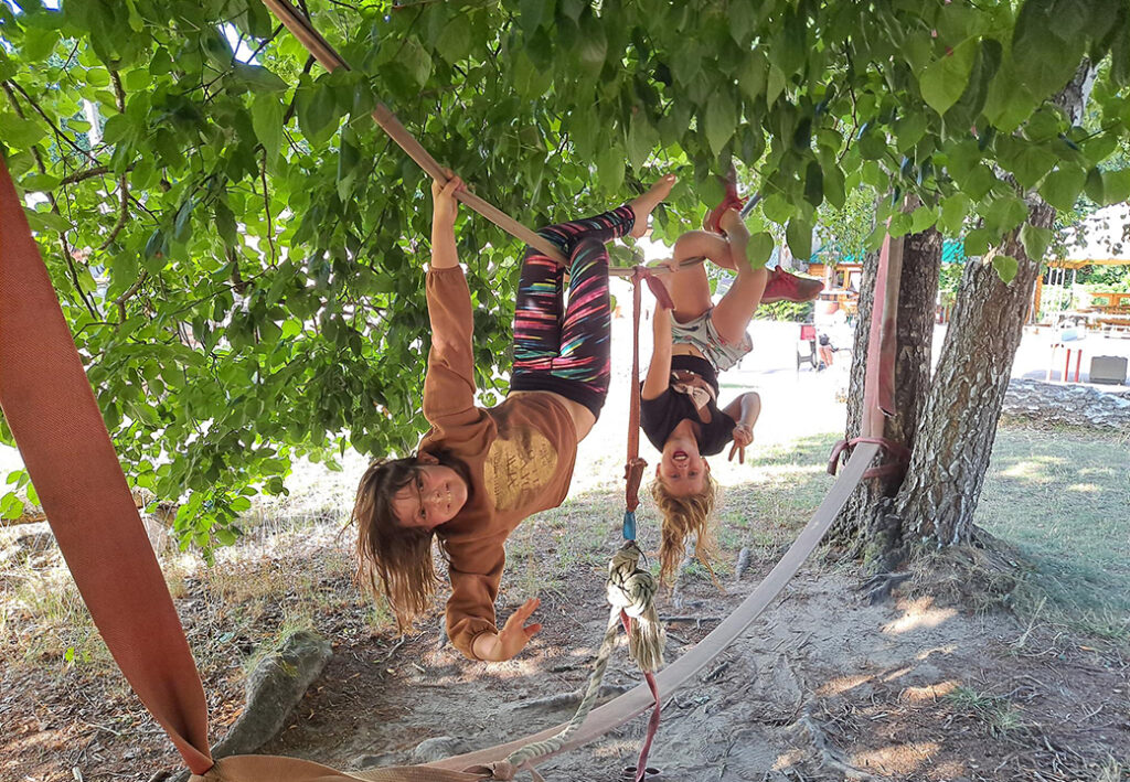 Zwei Kinder h&auml;ngen kopf&uuml;ber auf einem Slackline-Rigg, umgeben von B&auml;umen.