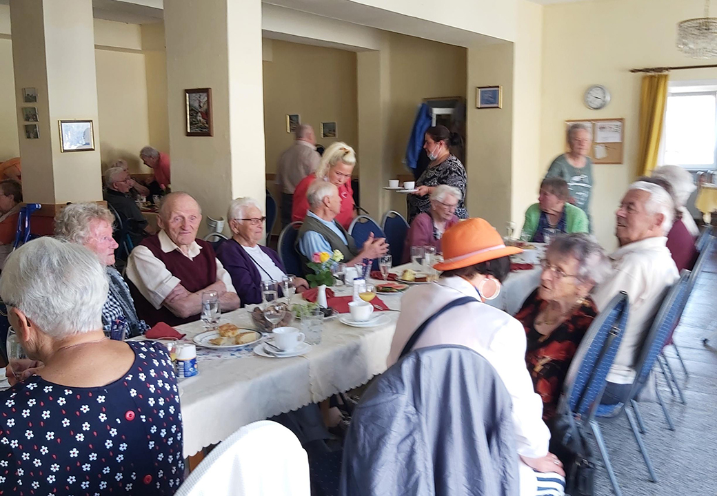 Eine Versammlung von älteren Menschen in einem Gemeinschaftsraum, die an einem langen Tisch sitzen und speisen.