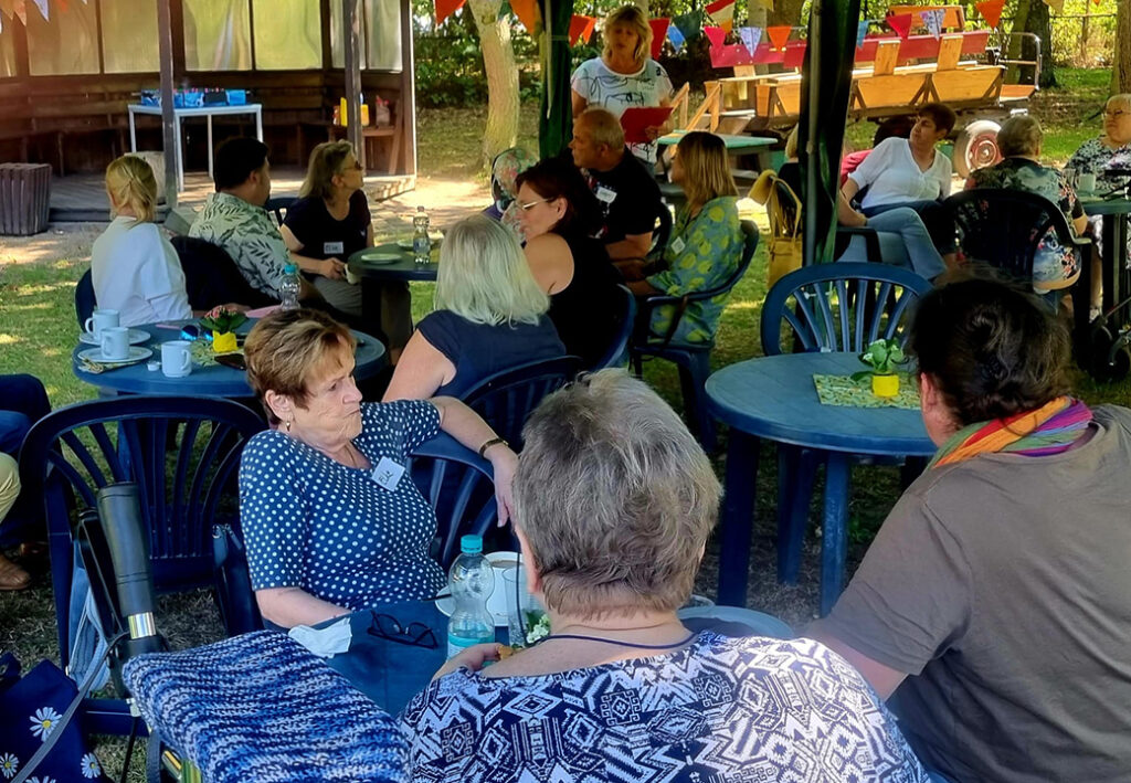 Eine Gruppe von Menschen sitzt in einem Garten mit Tisch und St&uuml;hlen.