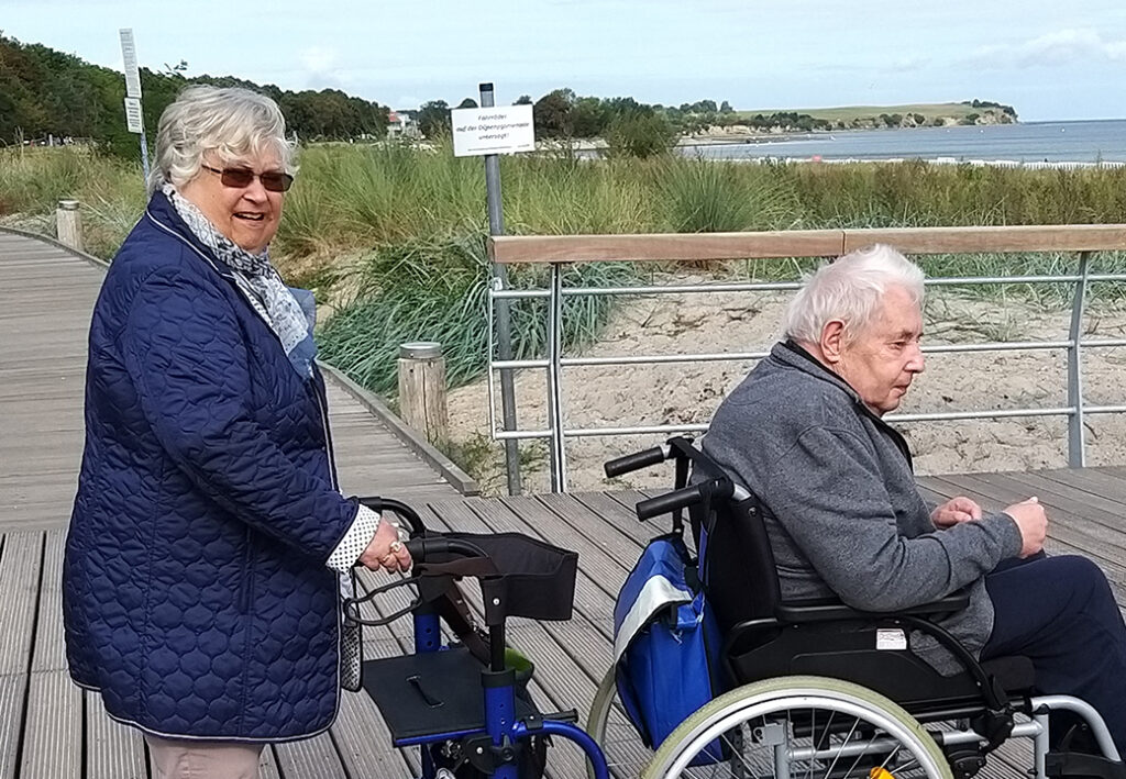 Zwei ältere Menschen auf einem Holzsteg am Strand, eine Person in einem Rollstuhl, die andere schiebt.