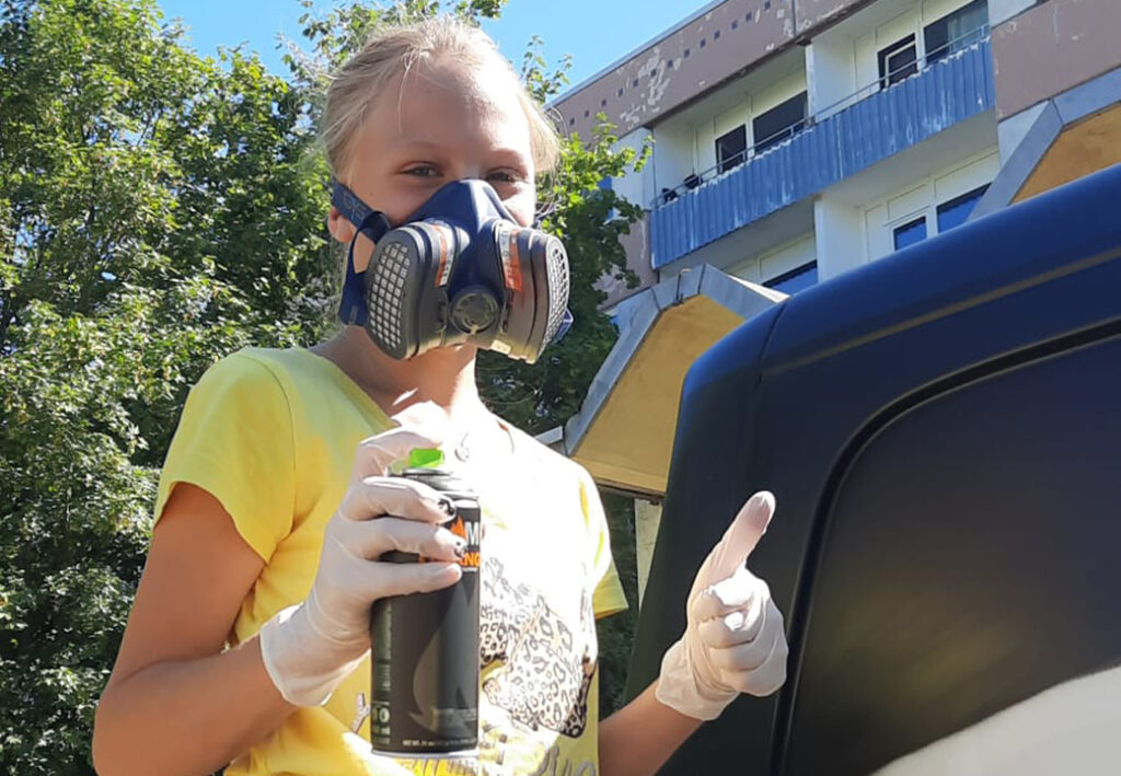 Eine Person mit Atemschutzmaske und Handschuhen h&auml;lt eine Spr&uuml;hdose und zeigt einen Daumen hoch vor einem Geb&auml;ude.