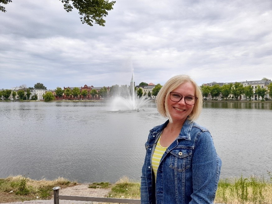 Eine Frau in einer Jeansjacke steht vor einem See mit einem Wasserbrunnen im Hintergrund.