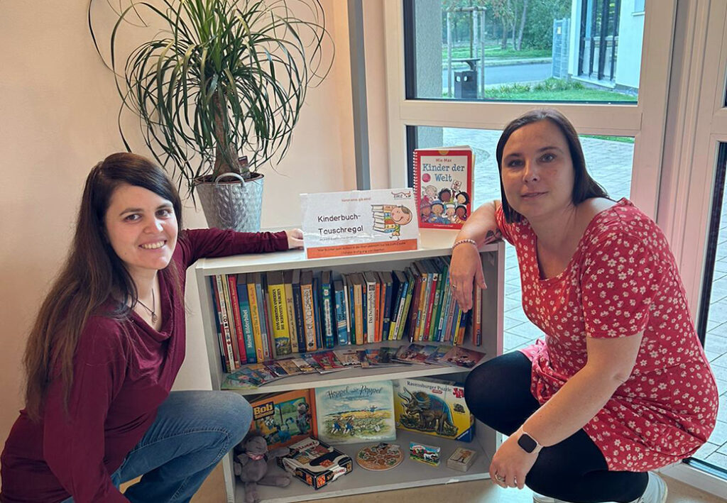 Zwei Frauen knien neben einem Kinderbuch-Tauschregal, das mit B&uuml;chern und Spielen gef&uuml;llt ist, in einem Raum mit Fenster und Topfpflanze.