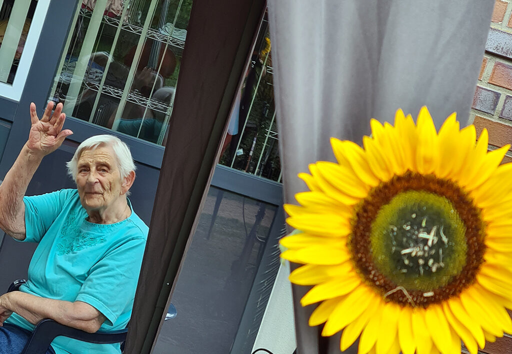 &Auml;ltere Frau winkt neben einer Sonnenblume.