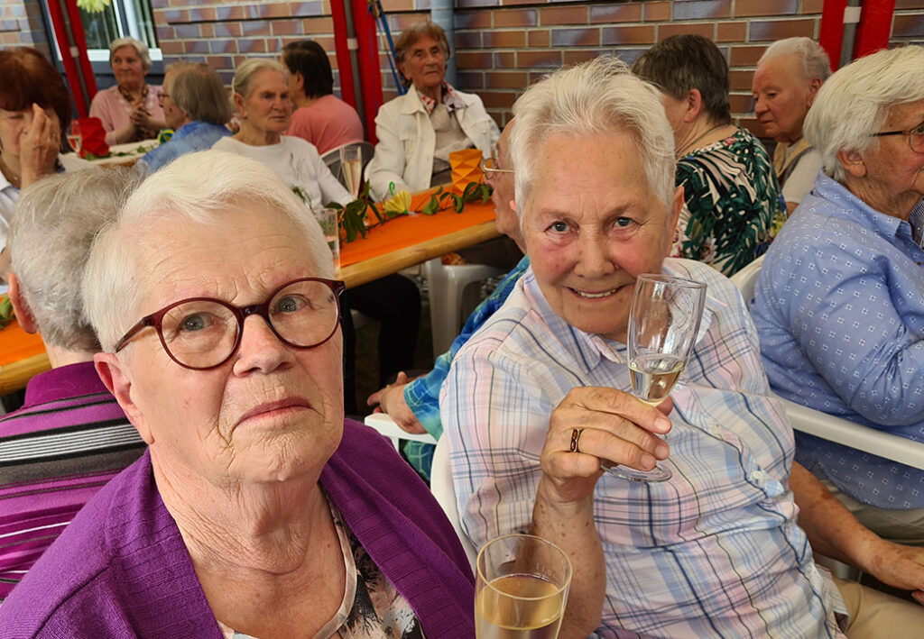 &Auml;ltere Damen bei einer Feier, die an einem Tisch mit dekorativer Tischdecke sitzen und Getr&auml;nke in der Hand halten.