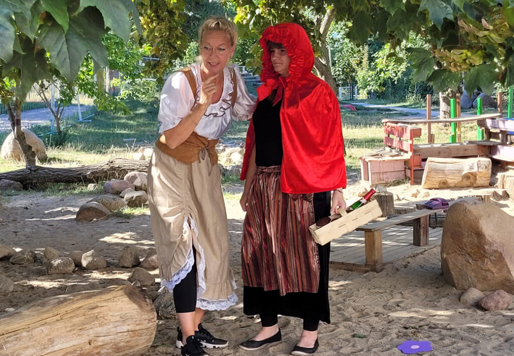 Zwei Personen, eine als Großmutter und die andere als Rotkäppchen verkleidet, stehen im Freien unter einem Baum.
