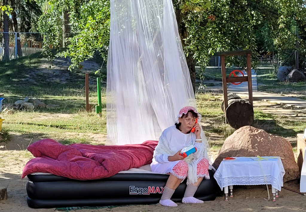 Eine Person sitzt auf einem schwarzen Luftbett im Freien, umgeben von Natur. Sie tr&auml;gt Schlafkleidung mit einem gepunkteten Helm und h&auml;lt ein rotes Spielzeugtelefon am Ohr. Ein transparenter Vorhang h&auml;ngt daneben, und auf einem Tisch liegen weitere Gegenst&auml;nde.