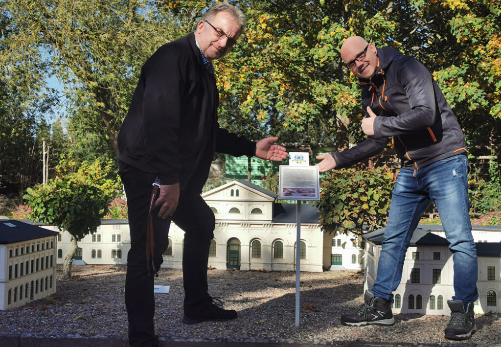 Zwei Männer vor einem Miniaturgebäude im Freien.