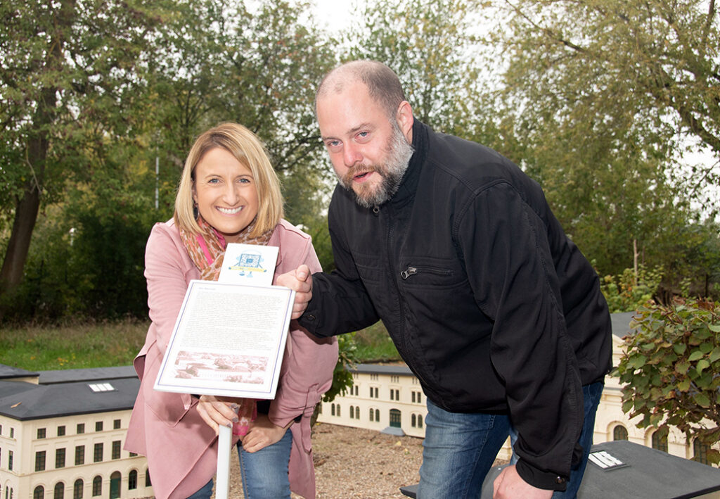 Eine Frau und ein Mann beugen sich l&auml;chelnd &uuml;ber ein Info-Schild in einer Parklandschaft.