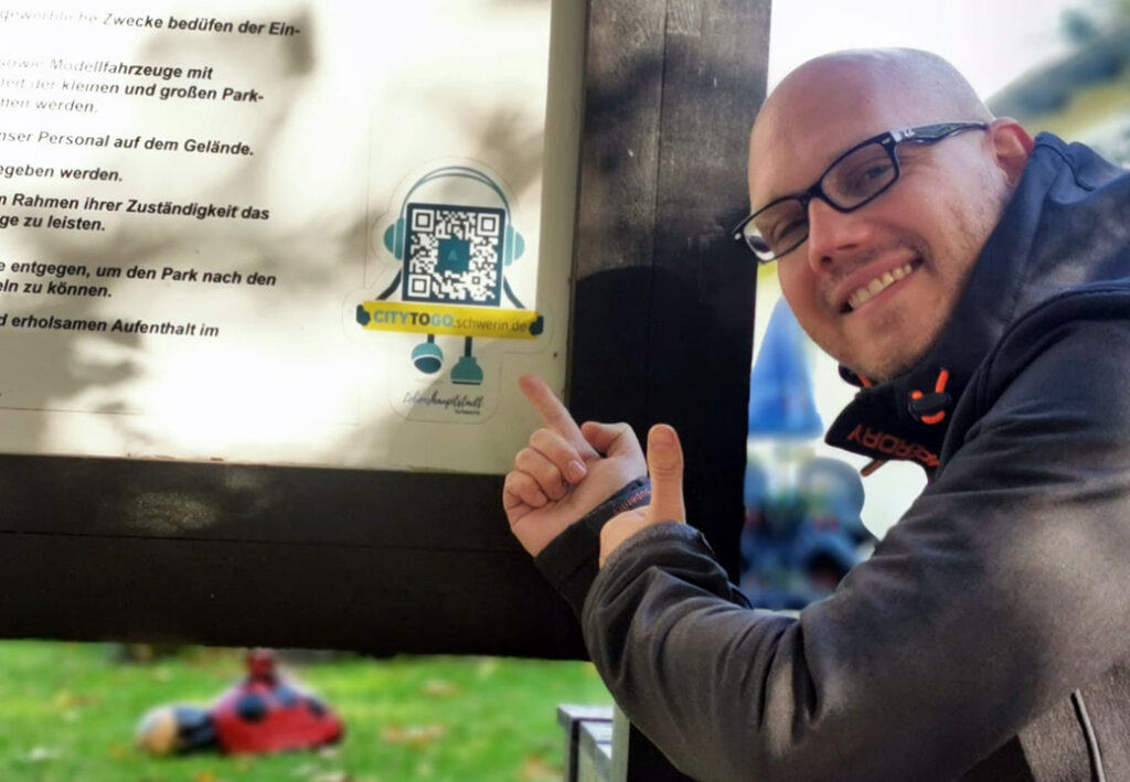 Ein Mann zeigt l&auml;chelnd auf einen QR-Code auf einem Schild.