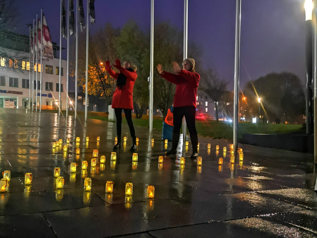 Zwei Personen in roten M&auml;nteln stehen bei Einbruch der Dunkelheit im Freien, umgeben von vielen leuchtenden Laternen.