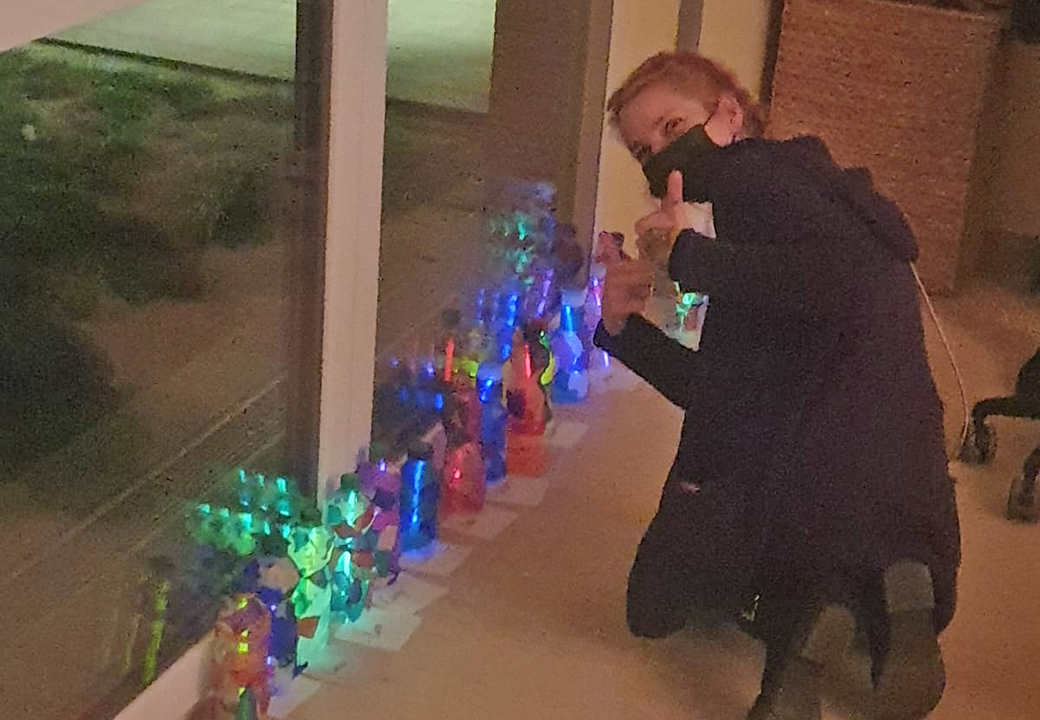 Person kniet neben einer Reihe von bunt beleuchteten Flaschen an einem Fenster