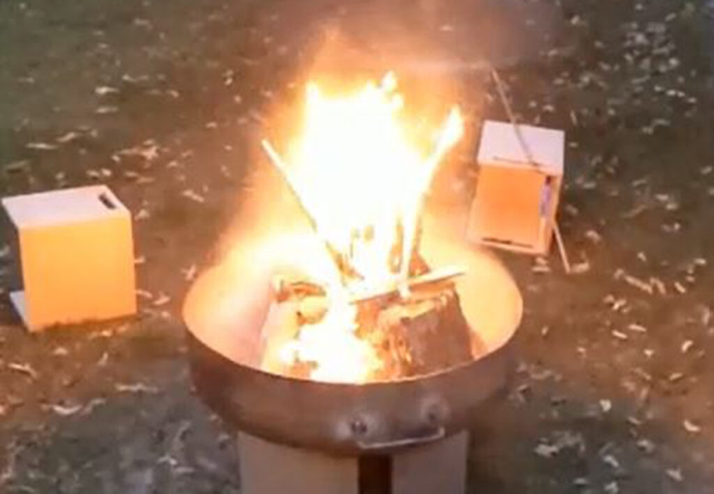 Feuerstelle mit Flammen in einer Metallfeuerschale, umgeben von niedrigen Holzhockern auf grasbewachsenem Boden.