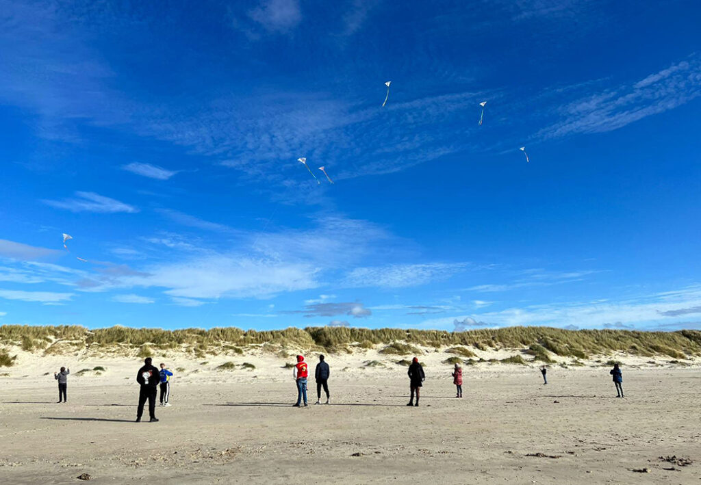 Personen am Strand mit fliegenden Drachen unter blauem Himmel.