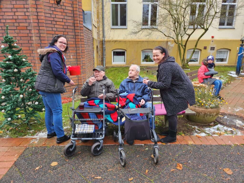 Zwei Frauen interagieren mit zwei &auml;lteren Herren vor einem Geb&auml;ude und einem Weihnachtsbaum.