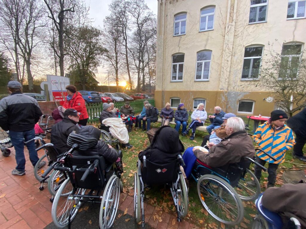 Eine Gruppe von Menschen, einige in Rollst&uuml;hlen, versammelt sich im Freien vor einem Geb&auml;ude mit mehreren Fenstern.