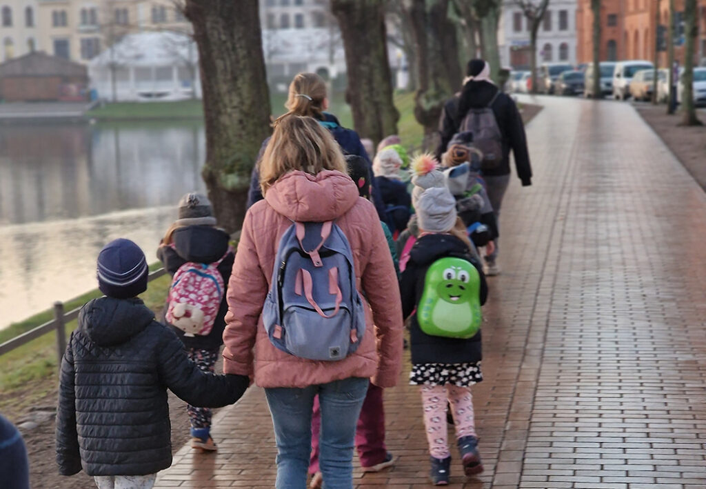 Eine Gruppe von Menschen, darunter Kinder mit bunten Rucks&auml;cken, die auf einem gepflasterten Gehweg entlang eines Gew&auml;ssers spazieren.