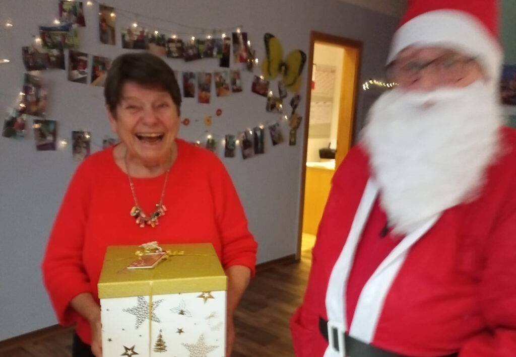 Eine lachende Frau h&auml;lt eine weihnachtlich verpackte Geschenkbox. Neben ihr steht eine Person in Weihnachtsmann-Kost&uuml;m.