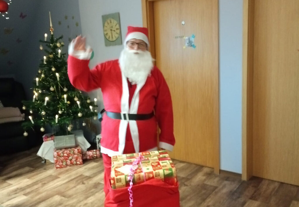 Eine Person in einem Weihnachtsmannkost&uuml;m steht neben einem geschm&uuml;ckten Weihnachtsbaum und einem Stapel Geschenke.