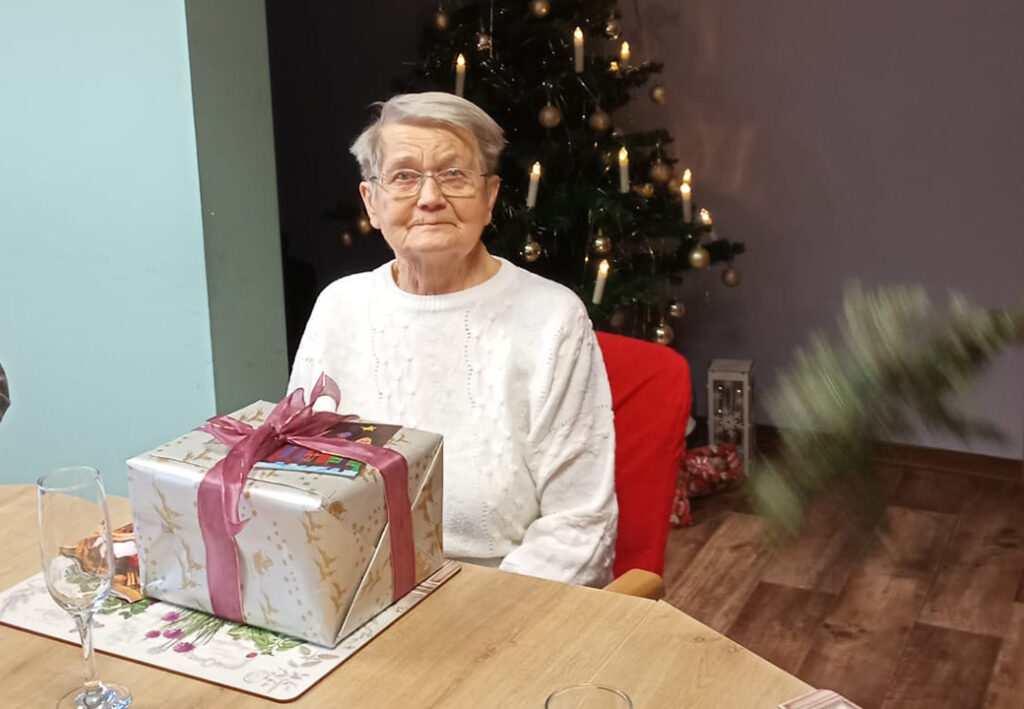 Eine &auml;ltere Frau sitzt an einem Tisch mit einem Geschenk vor einem Weihnachtsbaum.