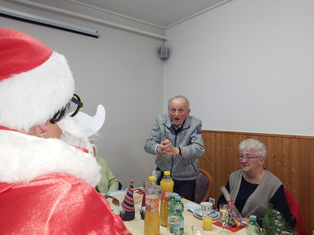 Ein &auml;lterer Mann spricht beeindruckt bei einer Feier, w&auml;hrend eine Frau und ein in Rot gekleideter Weihnachtsmann zusehen.