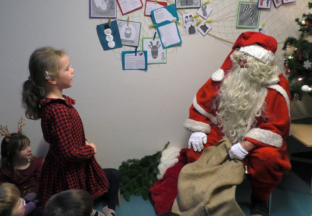 Bild mit einem sitzenden Weihnachtsmann und einem stehenden Kind in Winterkleidung.