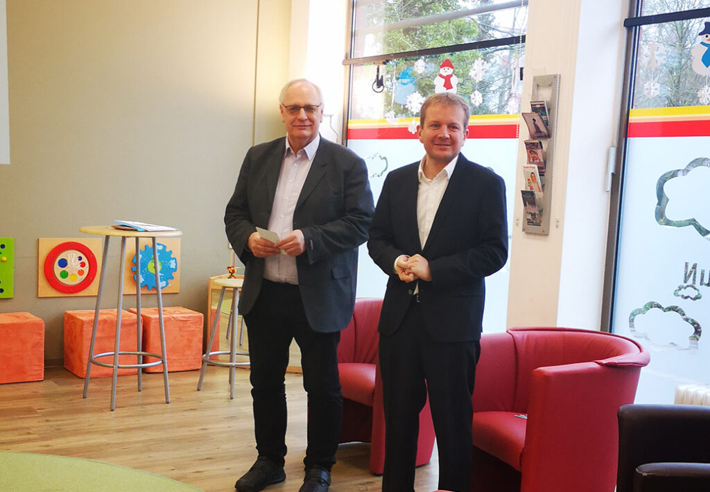 Zwei M&auml;nner stehen in einem offenen Raum mit roten St&uuml;hlen und bunten Kinderspieltischen.