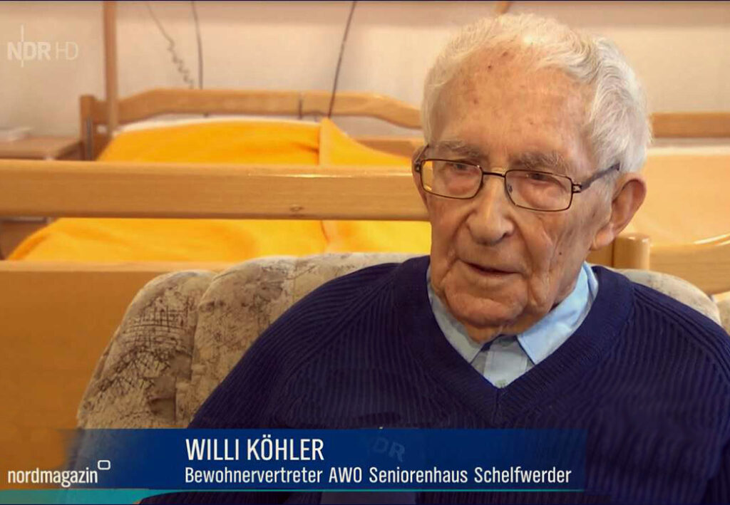 Ein &auml;lterer Herr mit Brille und blauem Pullover sitzt in einem Sessel vor einem Bett mit gelber Decke.