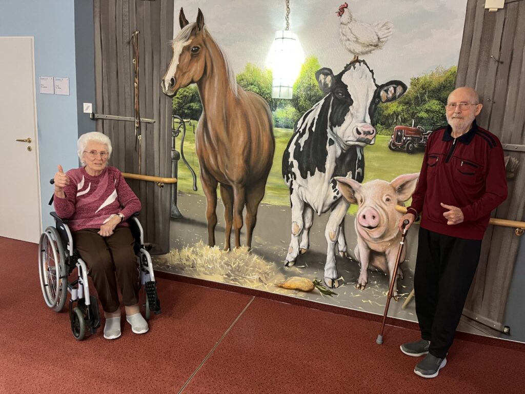 Zwei ältere Personen vor einem Wandbild mit Tieren in einem Innenraum