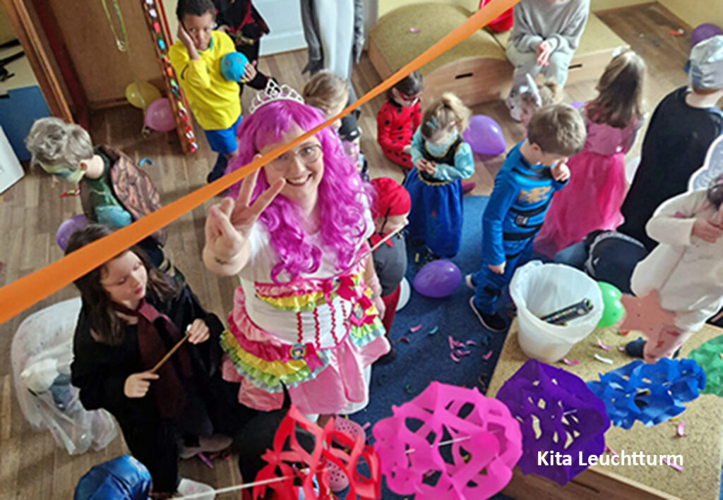 Kinder und Erzieherin in Kost&uuml;men bei einer Feier in einem Raum, dekoriert mit Luftballons und Girlanden.