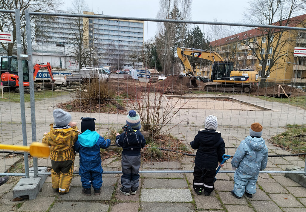 F&uuml;nf Kinder stehen vor einem Bauzaun und beobachten eine Baustelle.