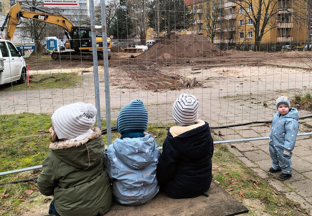 Drei Kinder sitzen vor einem Bauzaun, ein Kind steht daneben.