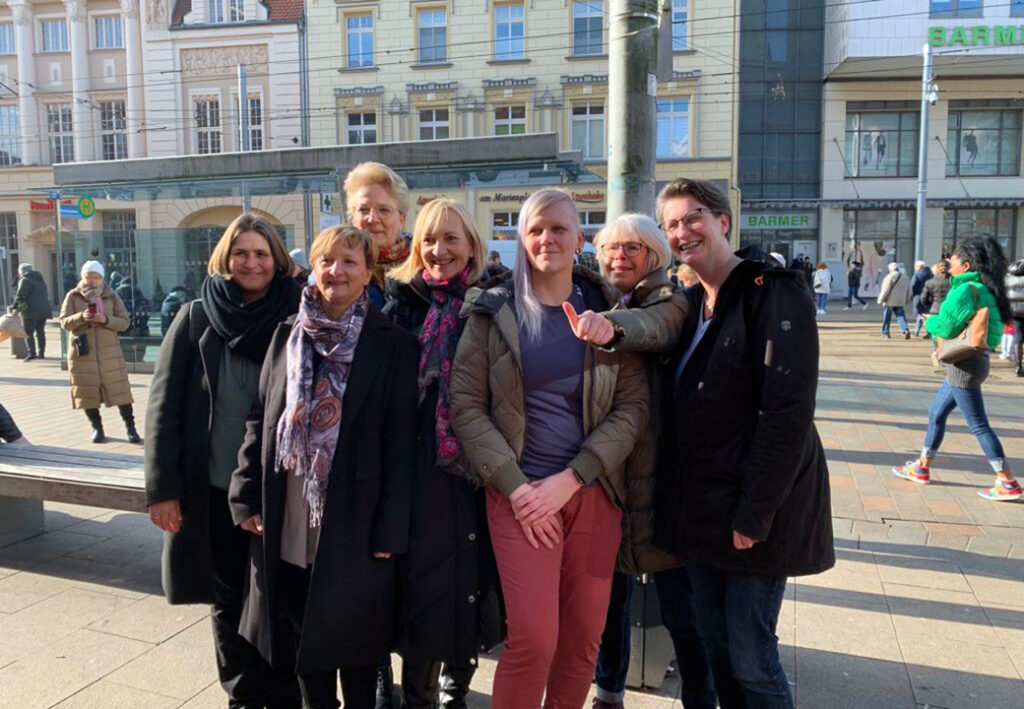 Eine Gruppe von sieben Frauen steht auf einem belebten Platz vor einem Geb&auml;ude.