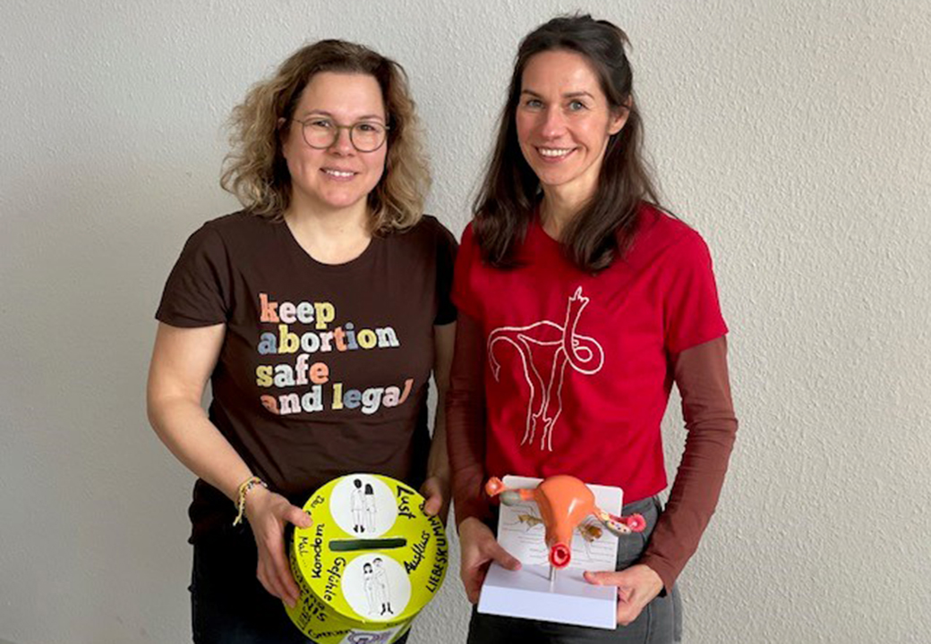 Zwei Frauen posieren lächelnd vor einer neutralen Wand.