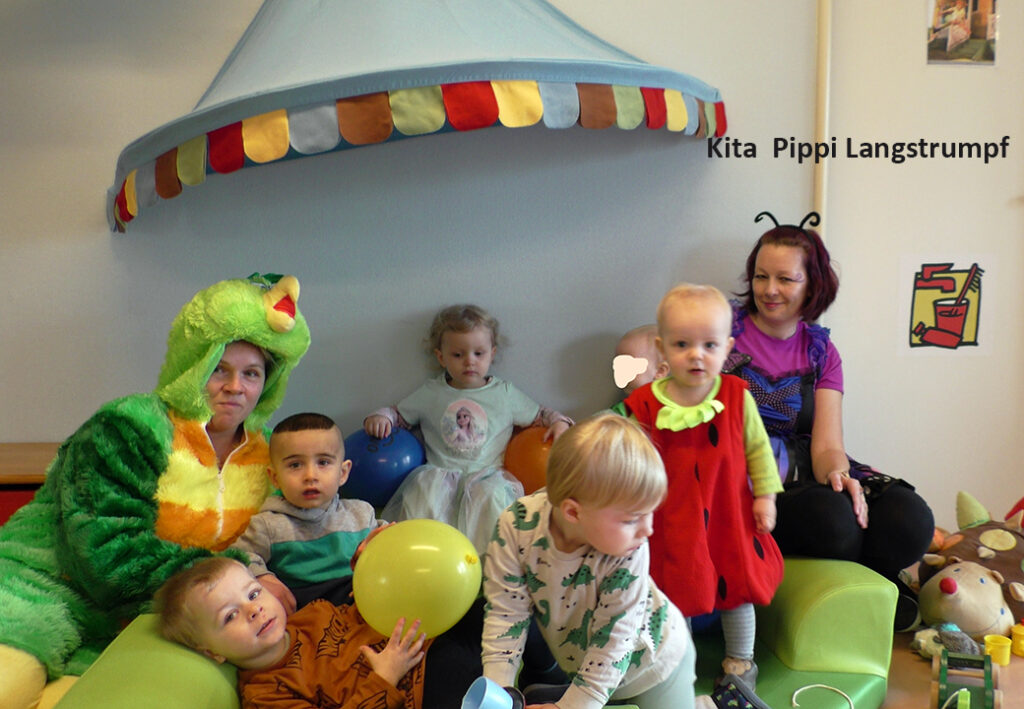 Gruppe von Kindern und Erwachsenen in Kost&uuml;men unter einem baldfach&auml;hnlichen Dach in einem Raum der Kita Pippi Langstrumpf.