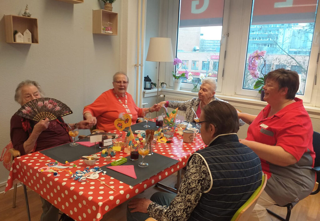 Eine Gruppe von f&uuml;nf Personen, darunter &auml;ltere Damen, sitzen fr&ouml;hlich an einem Tisch in einem gem&uuml;tlichen Raum. Der Tisch ist bunt dekoriert mit Masken und Gl&auml;sern.