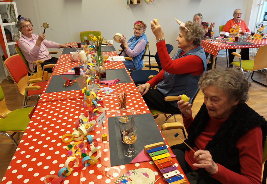 Eine Gruppe &auml;lterer Menschen sitzt an einem mit rotem, gepunktetem Tischtuch gedeckten Tisch und h&auml;lt Musikinstrumente.