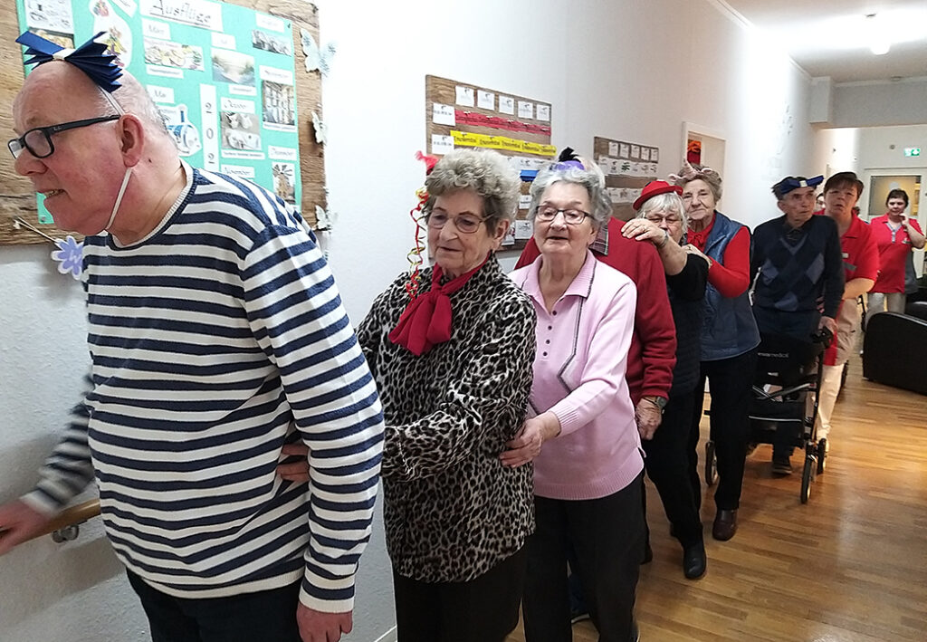 Eine Gruppe &auml;lterer Menschen tanzt im G&auml;nsemarsch in einem Gang eines Pflegeheims.