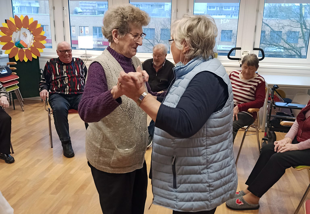 Zwei ältere Frauen tanzen in einem Gemeinschaftsraum, umgeben von sitzenden Senioren.