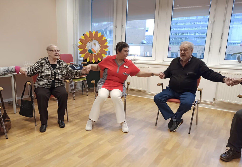 Drei Personen halten sich an den H&auml;nden und sitzen im Kreis auf St&uuml;hlen in einem Raum.