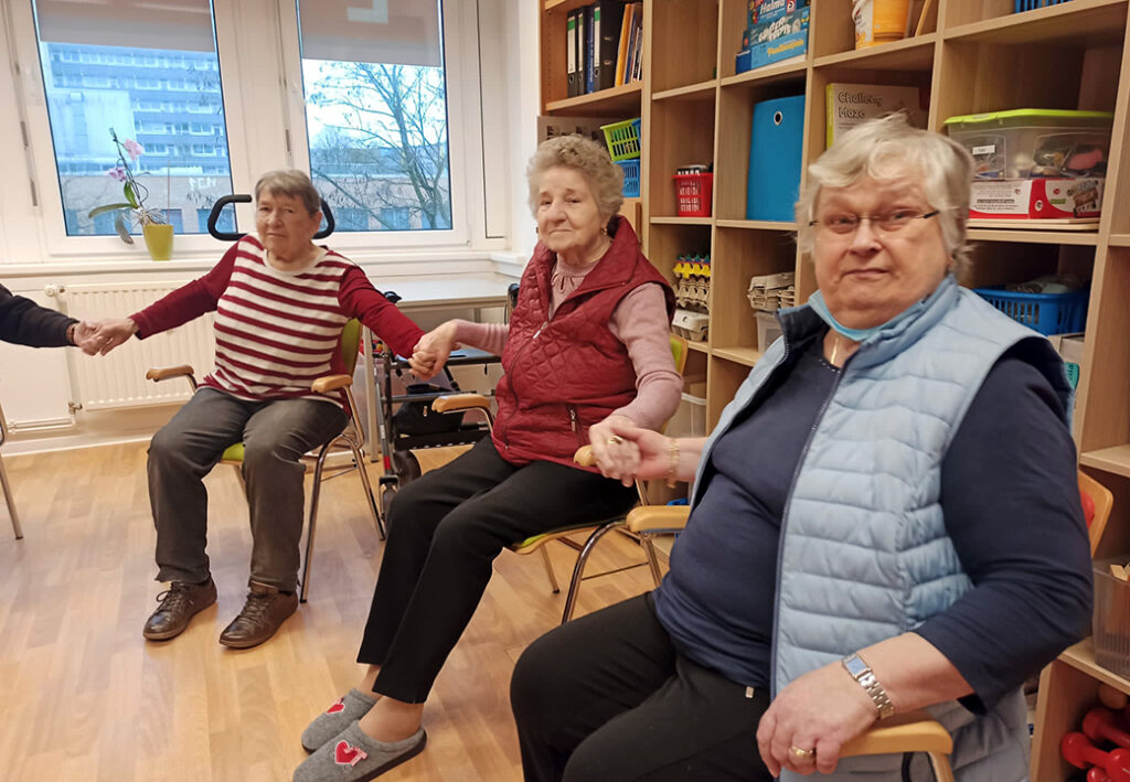 Drei &auml;ltere Frauen sitzen in einem Raum und halten sich an den H&auml;nden.
